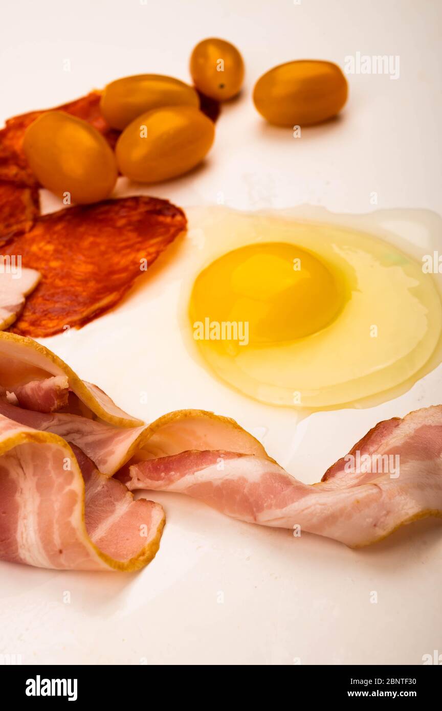 Ein zerbrochenes Hühnereier ohne Schale, Scheiben Wurst und Speck und Tomaten auf weißem Hintergrund. Nahaufnahme Stockfoto