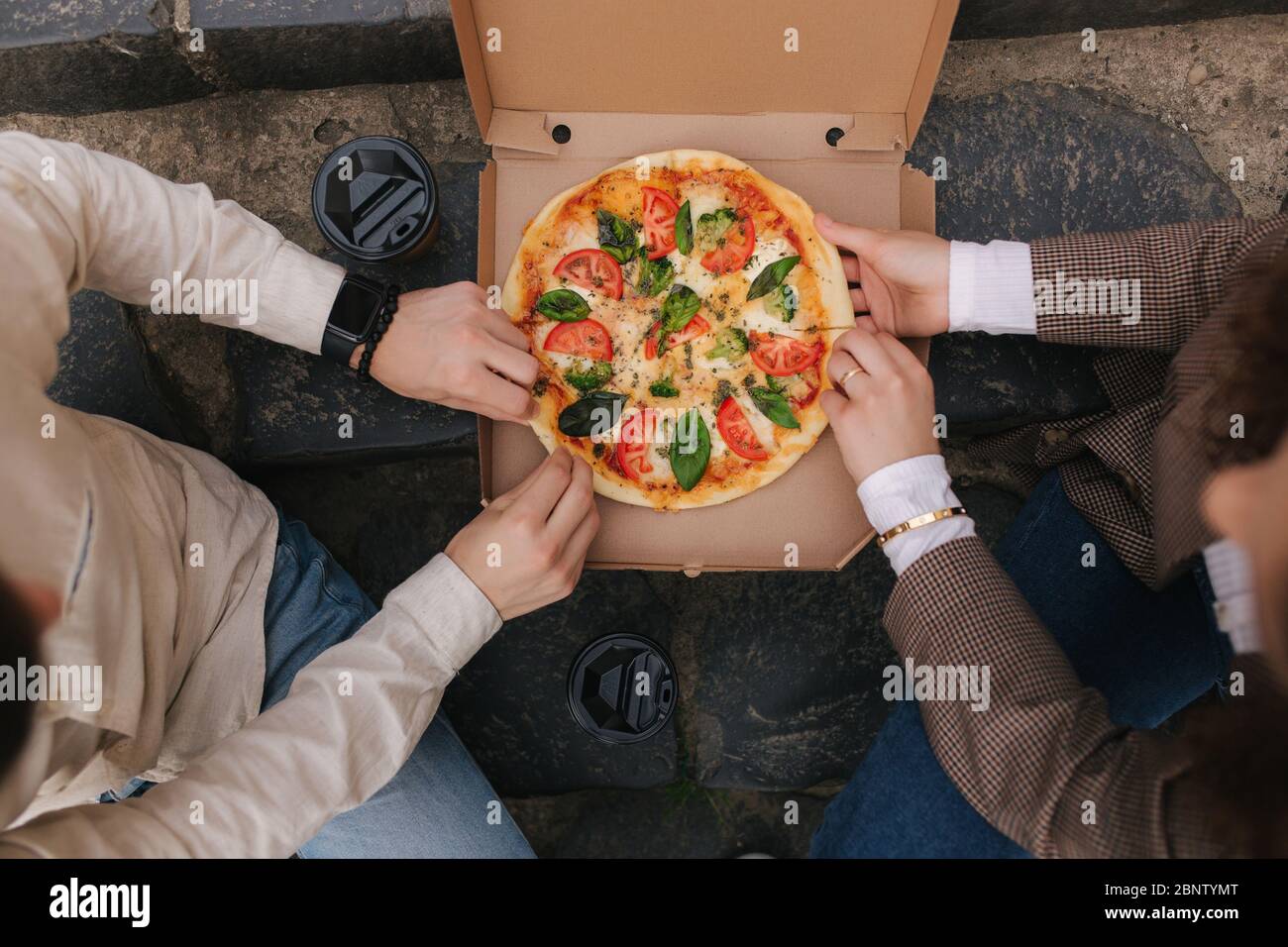 Draufsicht Bild von paar Schnapp Scheiben Pizza aus der Box im Freien. Mann und Frau Hände, die Pizza nehmen. Vegane Pizza mit frischen Tomaten Basilikum und Stockfoto