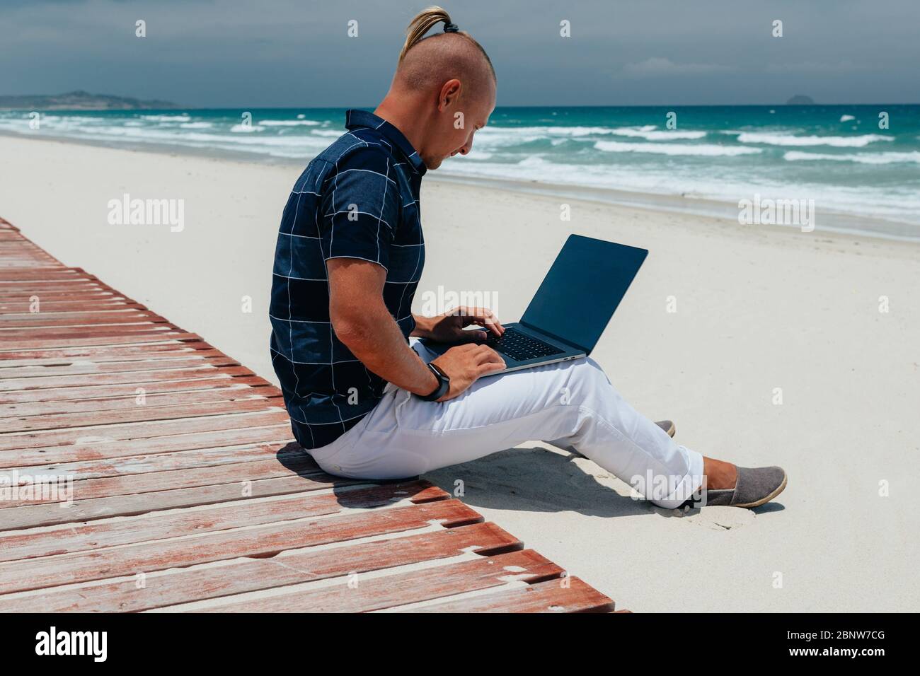 Junger Mann, der mit einem Laptop arbeitet und am Strand sitzt. Geschäftsmann, der an Remote-Arbeit auf einer Reise arbeitet. Blogger, Freiberufler macht Stockfoto