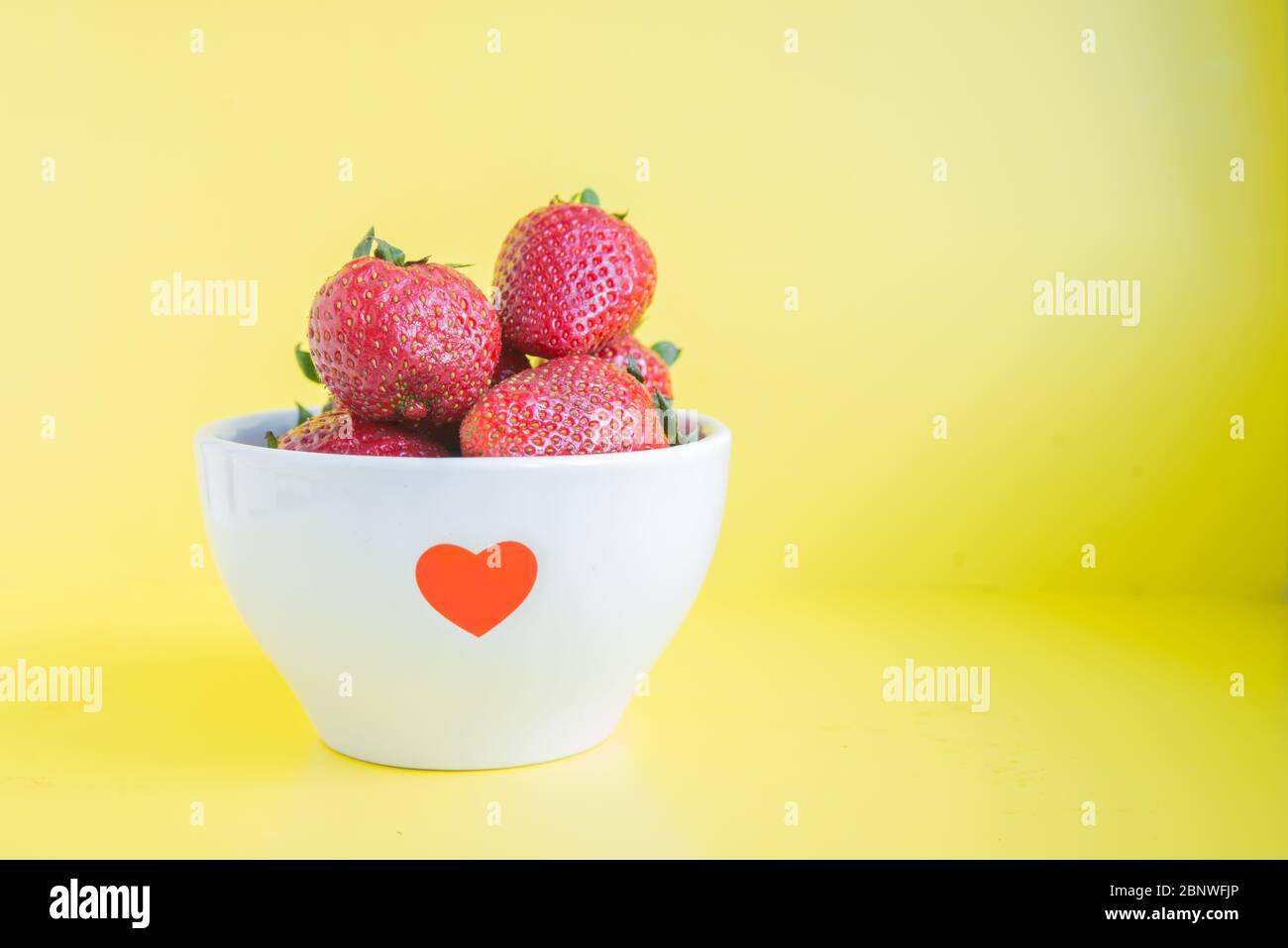 Frische Erdbeeren in weißer Schüssel mit rotem Herz auf gelbem Hintergrund mit Copyspace Stockfoto