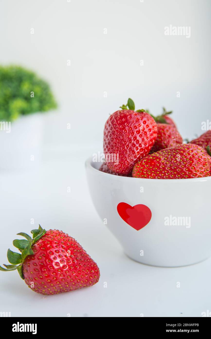 Frische Erdbeeren in weißer Schüssel mit rotem Herz auf weißem Hintergrund mit Copyspace vertikales Foto Stockfoto