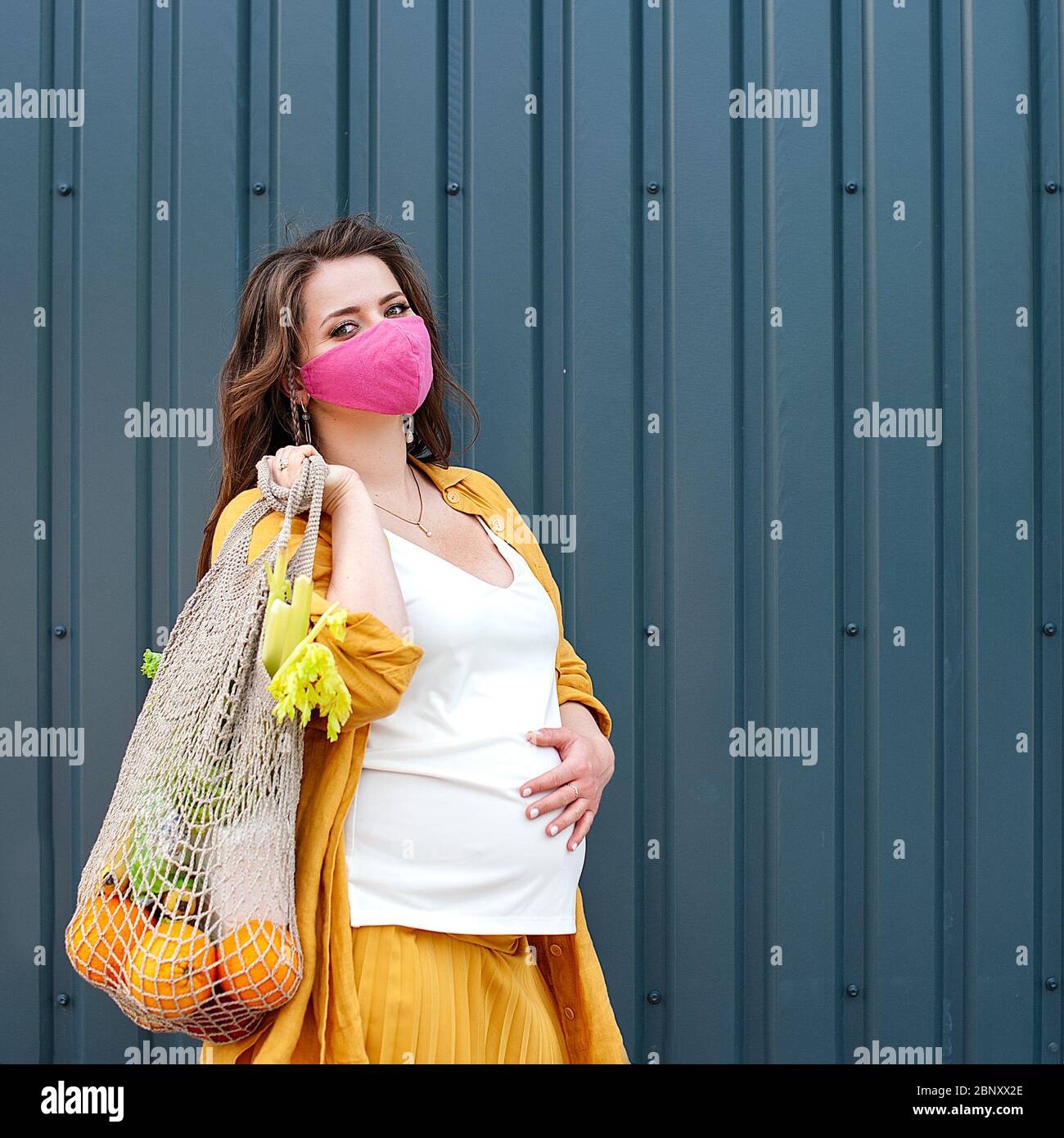 Schwangere Frau in einer Schutzmaske und gelbe Kleidung mit einem Öko-Beutel mit Obst und Gemüse auf einem blauen Wandhintergrund. Kunststoff-freies Konzept Sust Stockfoto