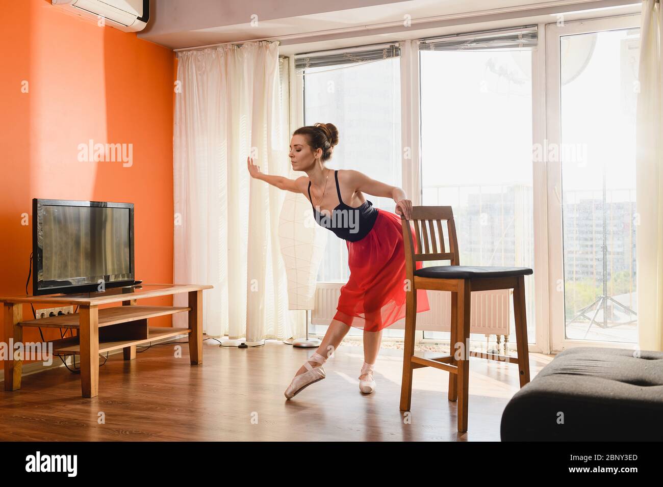 Schöne Ballerina Stretch unten Hand und Beine in beige spitzenschuhe, rot transparenten Rock, mit Haarbun, Stuhl halten, zu Hause lockdown Atmosphäre Stockfoto