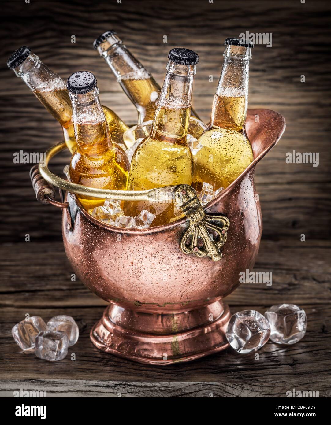 Viele Flaschen helles Bier mit Eisstücken befinden sich in einer alten Kupferschale. Hintergrund aus altem Holz Stockfoto