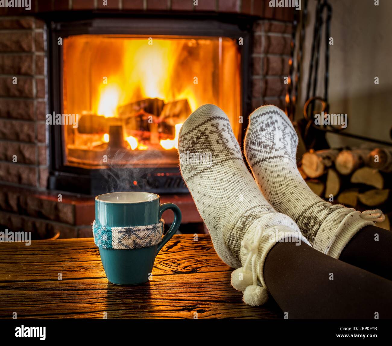 Weibliche Füße in weichen Wollsocken und brennendes Feuer im Kamin im Hintergrund. Das Aufwärmen und Abwickeln in der Nähe des Kamins mit einer Tasse heißem Getränk ist das sein Stockfoto