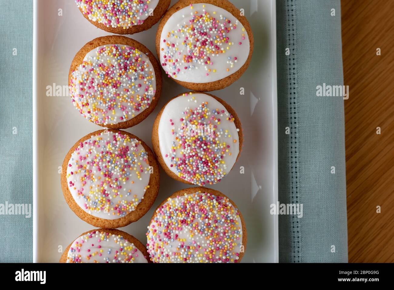 Iced Märchen Kuchen in einer Reihe auf einem langen weißen Teller sitzen auf einem blauen Stoff. Die Feigkuchen sind mit bunten Zuckerstreuseln bedeckt. Stockfoto