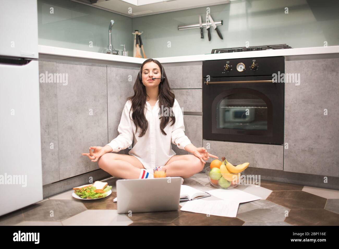 Schöne schwarzhaarige Frau arbeitet von zu Hause aus. Ein weibliches Model sitzt in einer Yoga-Pose auf dem Küchenboden. Das Ende einer ausgewogenen und ruhigen Arbeit. Fühlt sich voll an Stockfoto