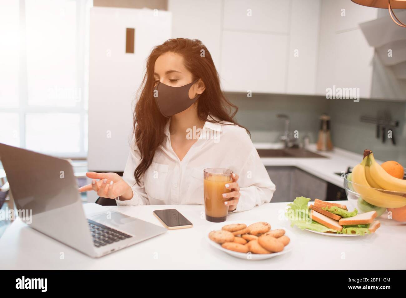 Wegen der Coronavirus-Pandemie, eine Frau in Isolation zu Hause. Sie arbeitet zu Hause, trägt eine Maske und hat eine Videokonferenz auf ihrem Laptop Stockfoto