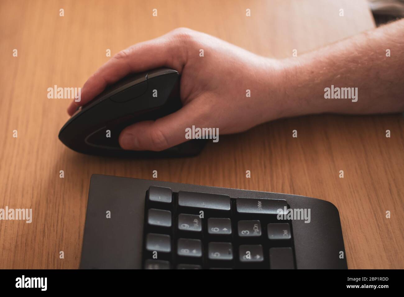 Ein Arm mit einer ergonomischen Maus auf einem Holztisch mit einer schwarzen Tastatur im Vordergrund Stockfoto