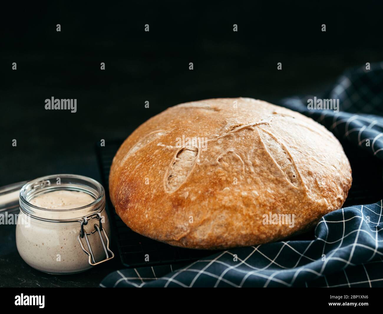 Weizenrunder Sauerteig Brot und Sauerteigstarter im Glasgefäß. Seitenansicht von köstlichen hausgemachten Sauerteig Brot auf schwarzem Hintergrund mit Kopierraum. Hausgemachtes Sauerteig Brot machen Konzept. Stockfoto