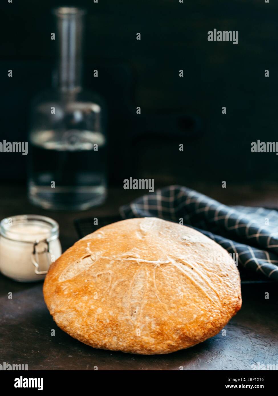 Weizenrunder Sauerteig Brot und Sauerteigstarter im Glasgefäß. Seitenansicht von köstlichen hausgemachten Sauerteig Brot auf schwarzem Hintergrund mit Kopierraum. Hausgemachtes Sauerteig Brot machen Konzept. Vertikal. Stockfoto