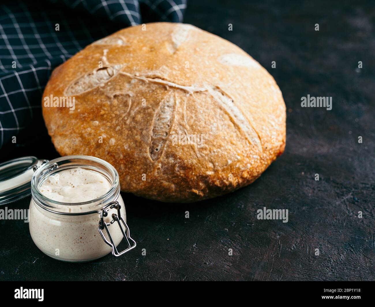 Sauerteig-Vorspeise und Weizensauerteig-Brot. Weizensaurer Teig Starter im Glas und köstliche hausgemachte runde Sauerteig Brot auf schwarzem Hintergrund, kopieren Raum. Hausgemachtes Sauerteig Brot machen Konzept Stockfoto