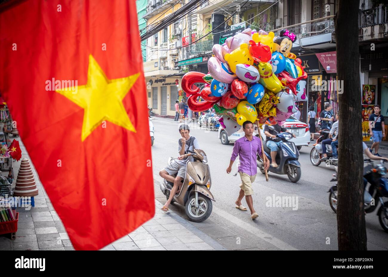 Hanoi, Vietnam - 30. April 2018: Straßenszene mit einem lokalen Händler, der einen Haufen bunter Ballons und die vietnamesische Nationalflagge im Vorschiff trägt Stockfoto