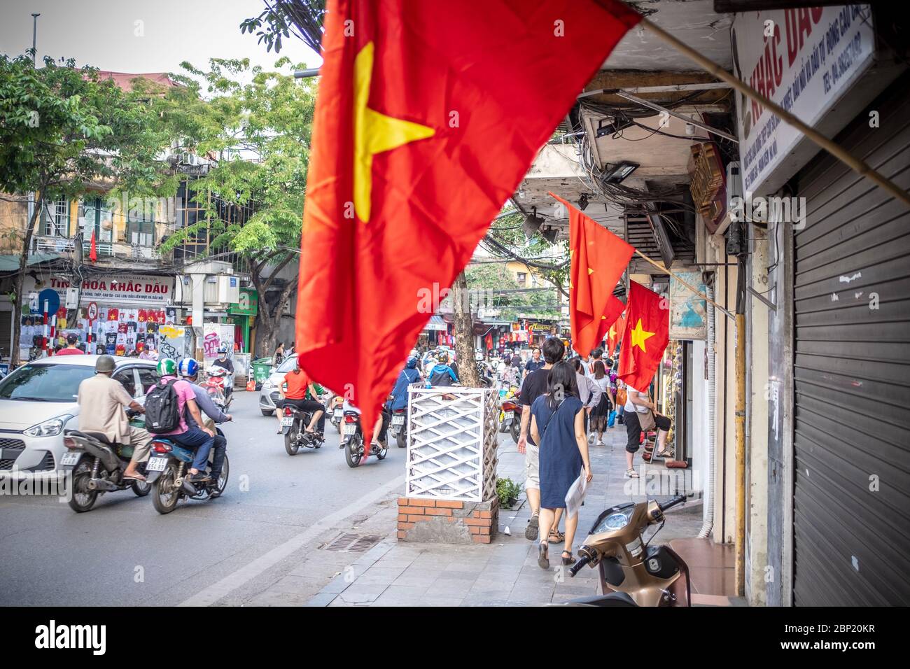Hanoi, Vietnam - 30. April 2018: Straßenszene mit Einheimischen, die ihre Fahrräder oder Motorräder auf einer belebten Straße mit vietnamesischer Nationalflagge befreien Stockfoto