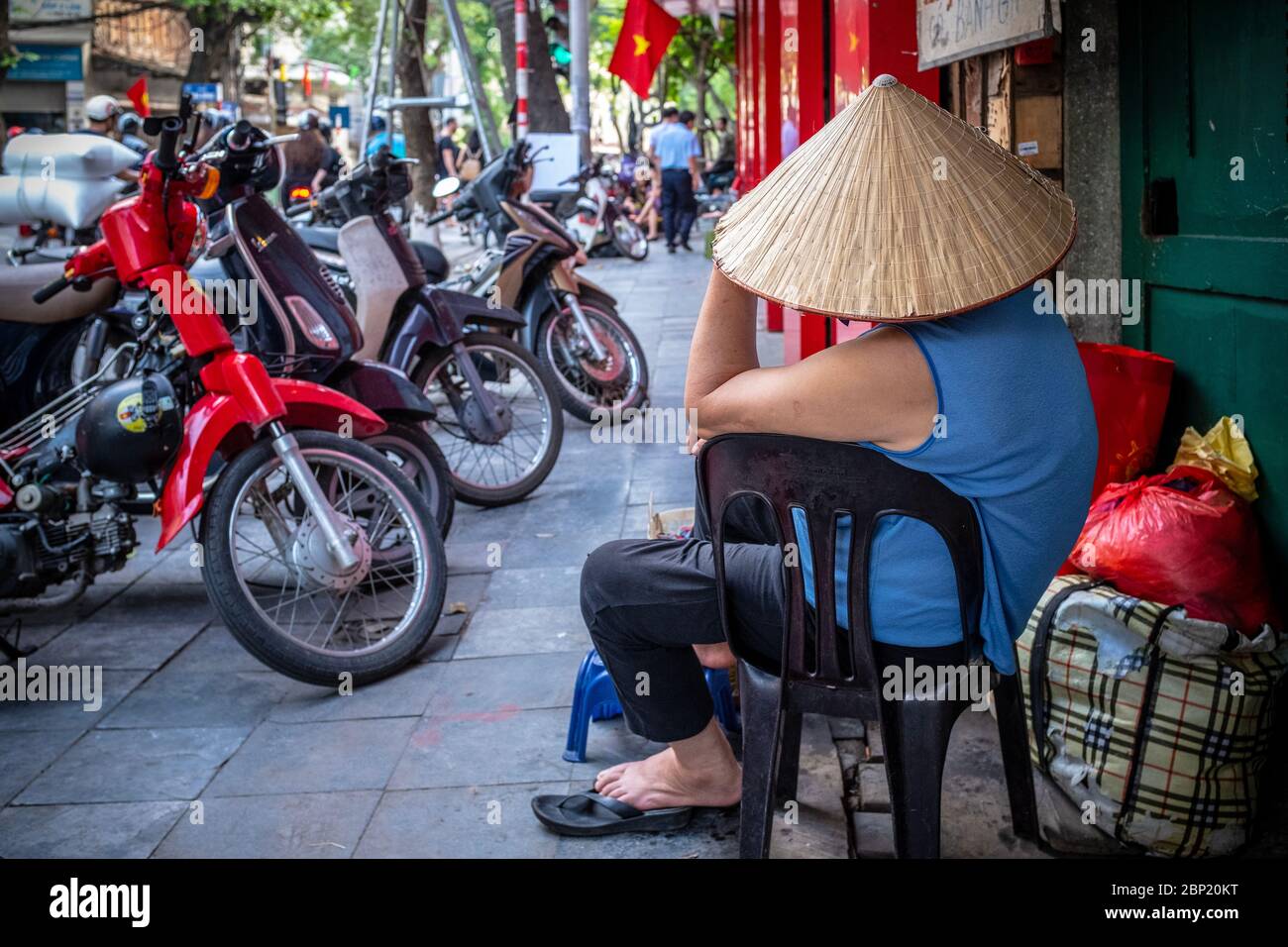 Hanoi, Vietnam - 30. April 2018: Straßenszene mit einem Mann, der einen Strohhut trägt und von hinten gesehen Motorräder auf dem Bürgersteig parkt Stockfoto