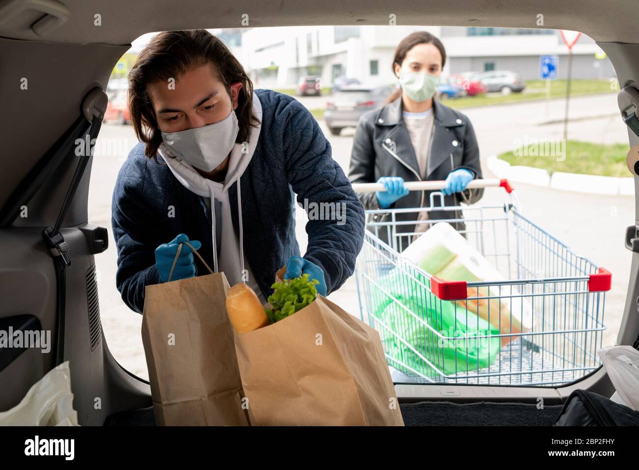 Junger Mann in Schutzhandschuhen und Maske, der Paperbags mit Lebensmitteln in den Kofferraum des Autos legt, während seine Frau mit einem Wagen in der Nähe steht Stockfoto