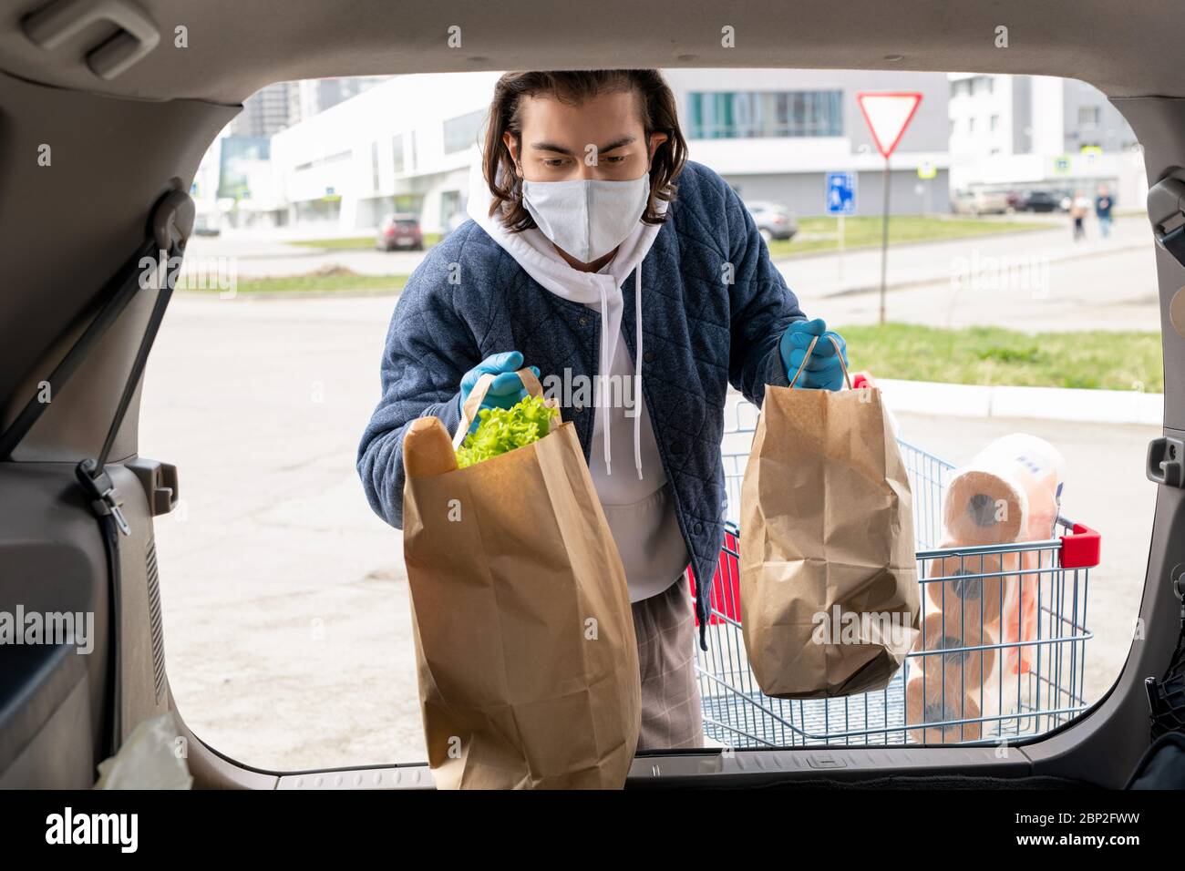 Junger Mann in Freizeitkleidung und Schutzhandschuhe und Maske stehen durch offenen Kofferraum beim Putting Paperbags mit Lebensmitteln im Inneren Stockfoto