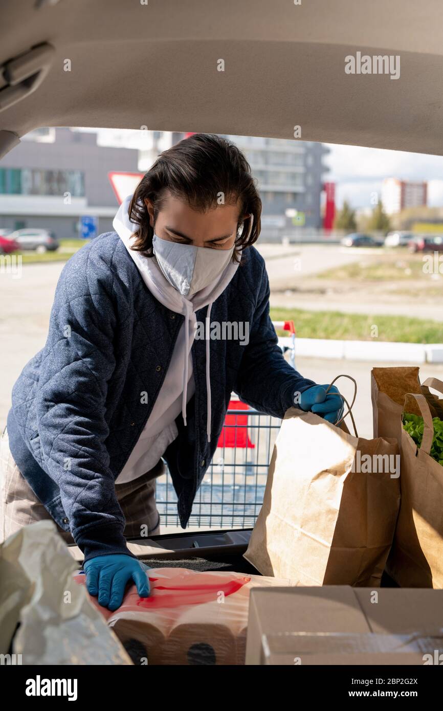 Junger Mann, der Paperbags mit frischen Lebensmitteln und Verpackung mit Toilettenpapier in den Kofferraum nach dem Besuch im Supermarkt Stockfoto