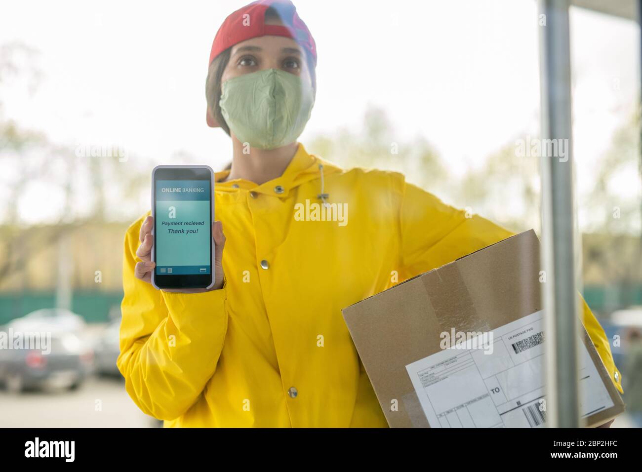 Junge Kurierin in Arbeitskleidung und Maske zeigt Transaktionsdaten auf dem Smartphone-Bildschirm, während auf den Kunden vor der Tür warten Stockfoto