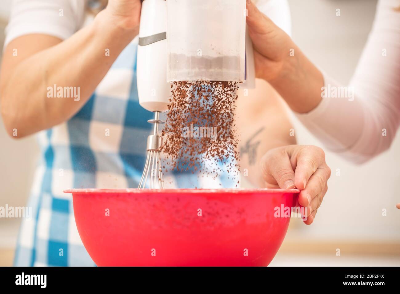 Detail auf der Hand von zwei Frauen, die Schokoladenpulver in eine rote Schüssel geben, mischen sie mit einem Handbesen. Stockfoto