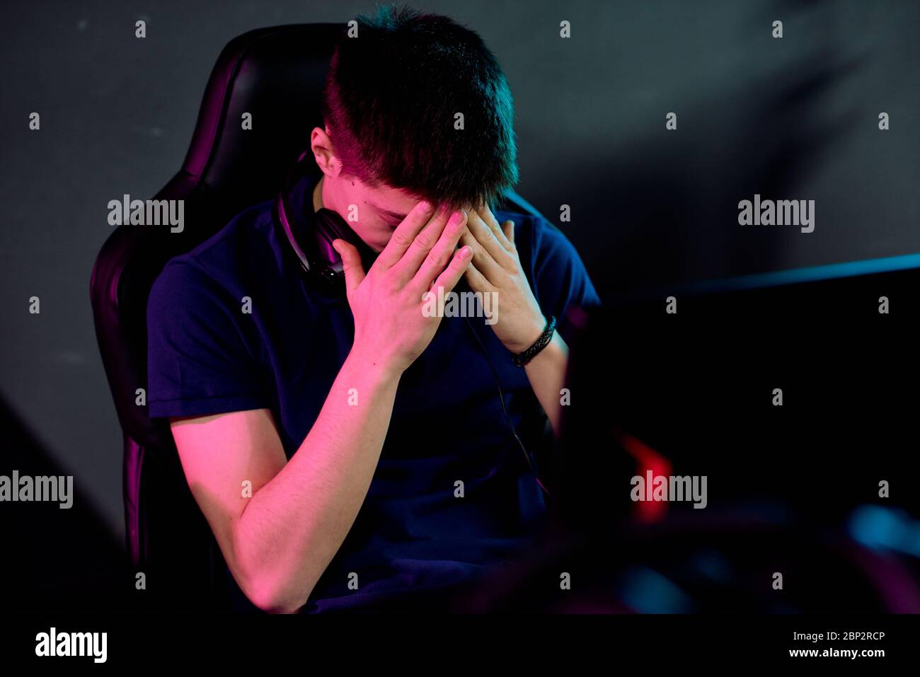 Besorgte junge Mann mit Kopfhörer um den Hals, die sein Gesicht mit den Händen während Cyber-Sport-Spiel während vor dem Monitor sitzen Stockfoto