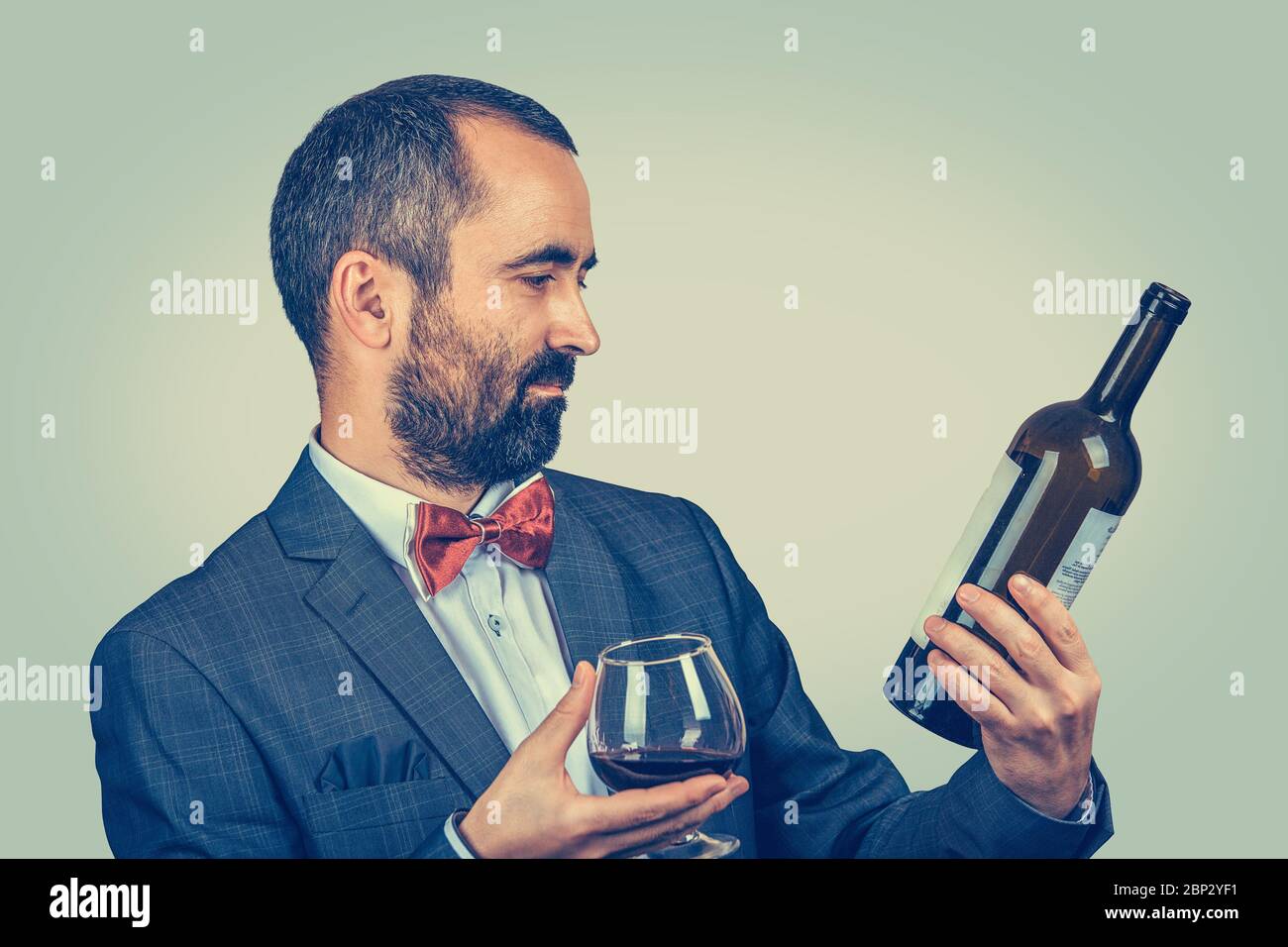 Ein Mann mit einem Glas Wein hält, schaut auf eine Flasche, lesen Sie die Informationen darüber. Gemischte Rasse bärtige Modell isoliert auf grün-gelbem Hintergrund Stockfoto