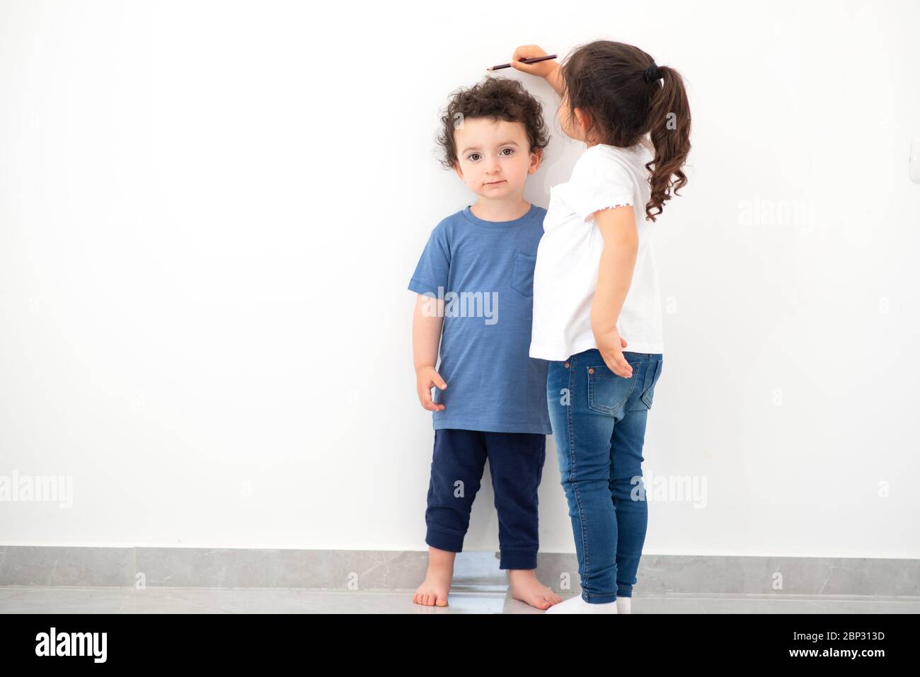 Nettes Mädchen misst Höhe eines lustigen Kindes mit lockigen Haaren. Schwester misst das Wachstum ihres Bruders an einer weißen Wand. Stockfoto