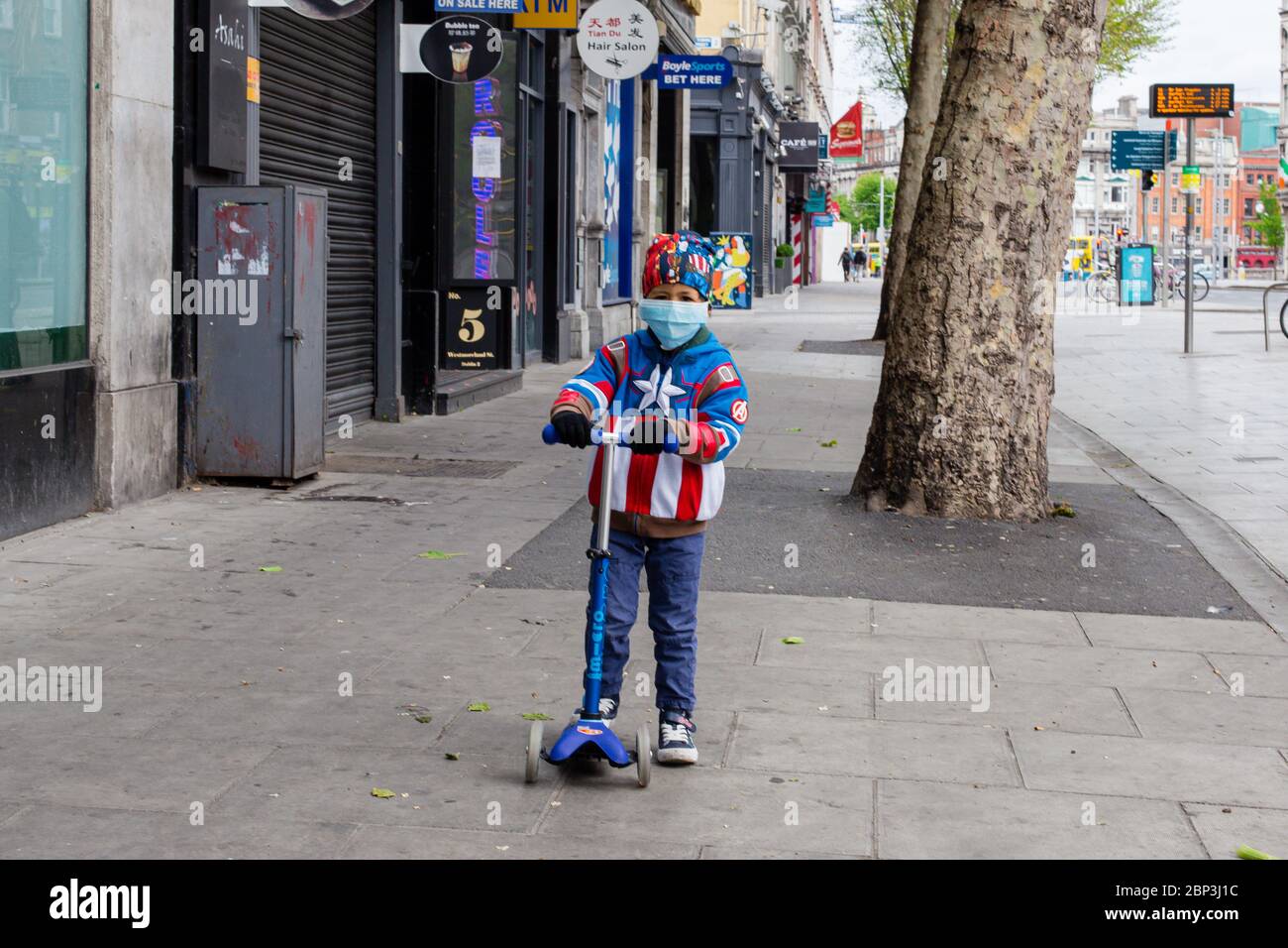 Junge auf einem Roller mit einer Superheldenjacke und einer schützenden Gesichtsmaske, die während der COVID-19-Pandemie durch das verlassene Westmoreland Str in Dublin scooting Stockfoto