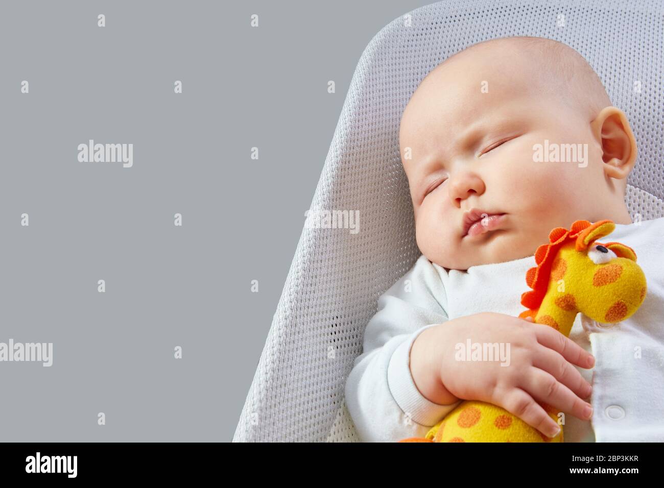 Baby schläft mit einer orangefarbenen Spielzeuggiraffe in einem Liegestuhl auf einem grauen Hintergrund mit Kopierraum. Stockfoto