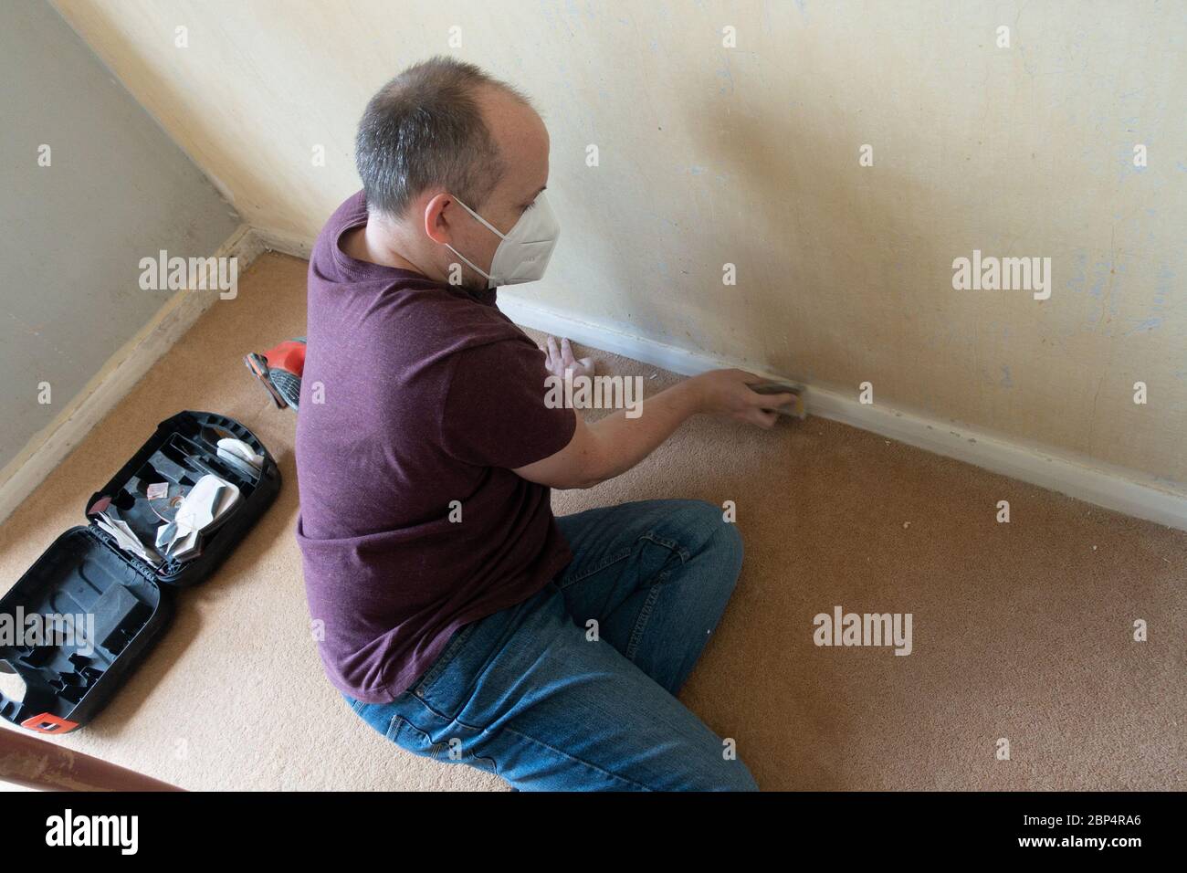 Ein Mann mittleren Alters, der die Sockelleiste vor dem Malen als Aprt eines DIY-Projekts abschleift. Stockfoto