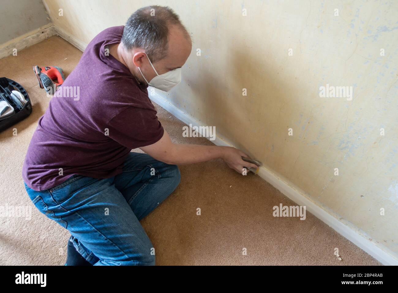 Ein Mann mittleren Alters, der die Sockelleiste vor dem Malen als Aprt eines DIY-Projekts abschleift. Stockfoto