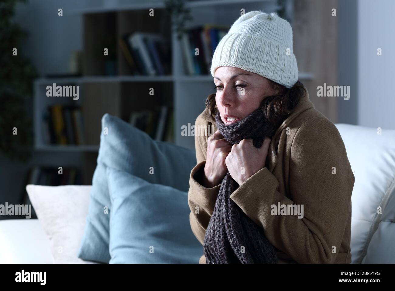 Kalte Erwachsene Frau mit Kleidung eiskalt sitzen auf dem Sofa in der Nacht zu Hause bedeckt Stockfoto