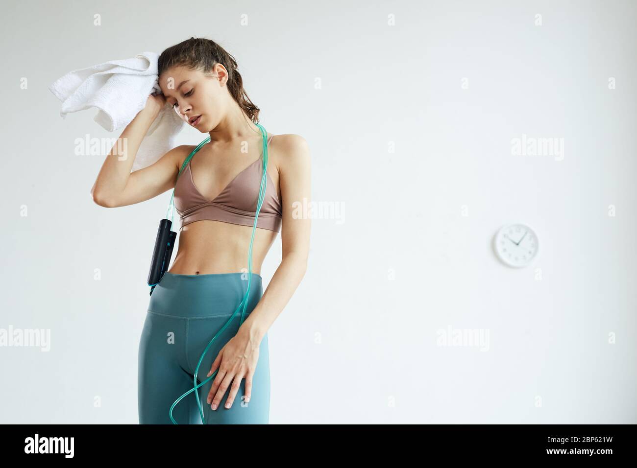 Minimales Taille-up-Porträt der erschöpften jungen Frau, die nach dem Fitness-Training Schweiß abwischt, steht gegen weiße Wand mit analoger Uhr, Kopierer-Raum Stockfoto