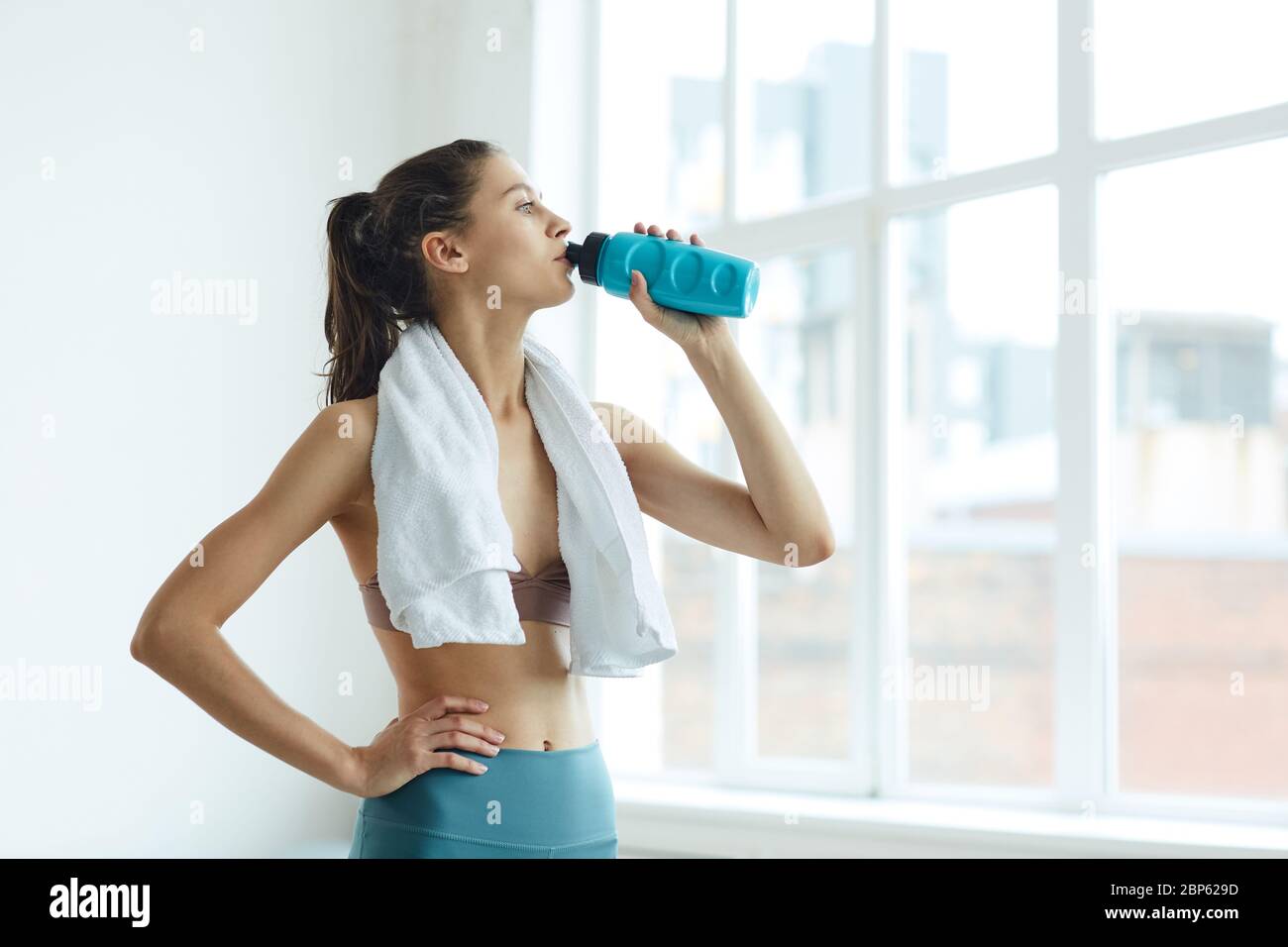 Minimal Taille oben Porträt von fit junge Frau Trinkwasser während am Fenster nach dem Sport-Training stehen, kopieren Raum Stockfoto