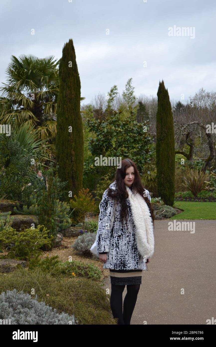 Brünette junge Dame mit langen Haaren und einem großen flauschigen Mantel und Schal stehen in einem formellen Garten an einem kalten Tag, vor Büschen und Bäumen. Stockfoto