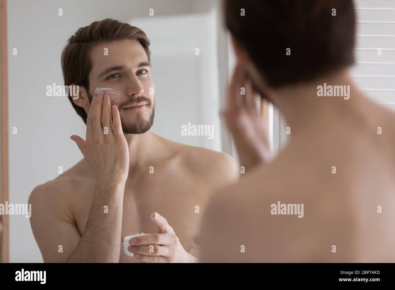 Junger Mann tragen feuchtigkeitsspendende Gesichtscreme im Badezimmer Stockfoto