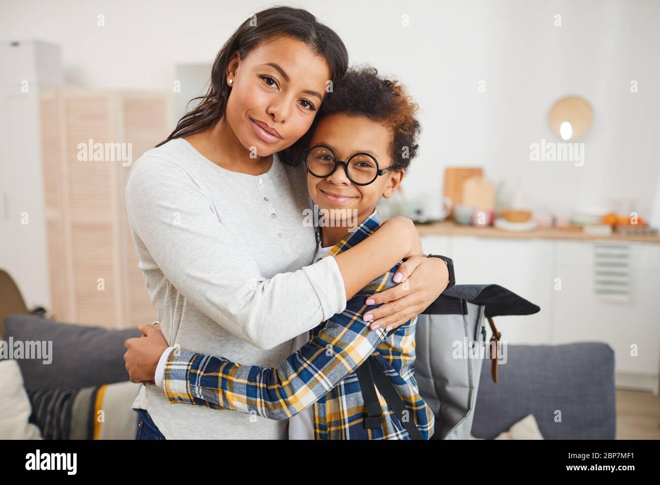 Taille-up-Porträt der glücklichen afroamerikanischen Mutter umarmt Sohn während er ihn in die Schule im September, beide Blick auf die Kamera Stockfoto