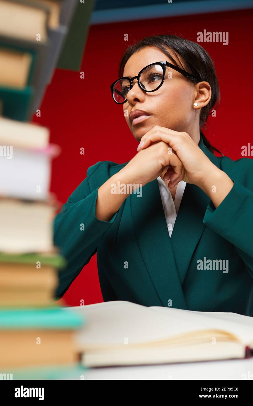 Niedrige Winkelansicht auf junge Lehrerin am Schreibtisch sitzend mit Stapel von Büchern im Vordergrund, auf rot geschossen Stockfoto
