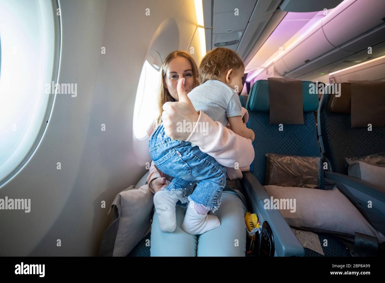 Konzept alles ist in Ordnung. Kleines niedliches Kleinkind springt auf den Knien mit einer jungen schönen Mutter in einem Flugzeug Stuhl. Mama zeigt Daumen nach oben und schaut an Stockfoto