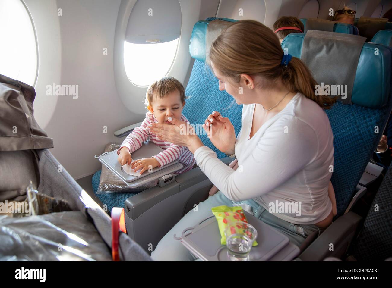 Kleines süßes Kleinkind Mädchen sitzt in einem Flugzeug in einem Stuhl am Bullauge, hält ein nasses Handtuch in den Händen und niest. Mama sitzt in der Nähe und bedeckt sie Stockfoto