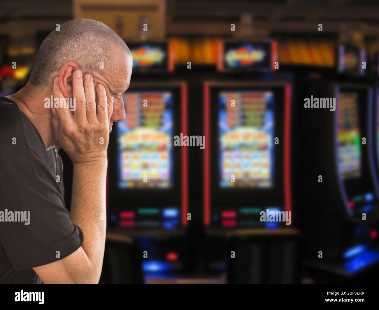 Trauriger und depressiver kaukasischer Mann, der mit den Händen auf dem Kopf vor Reihen von Casino-Spielautomaten sitzt. Spielsucht Thema Bild. Nahaufnahme Stockfoto