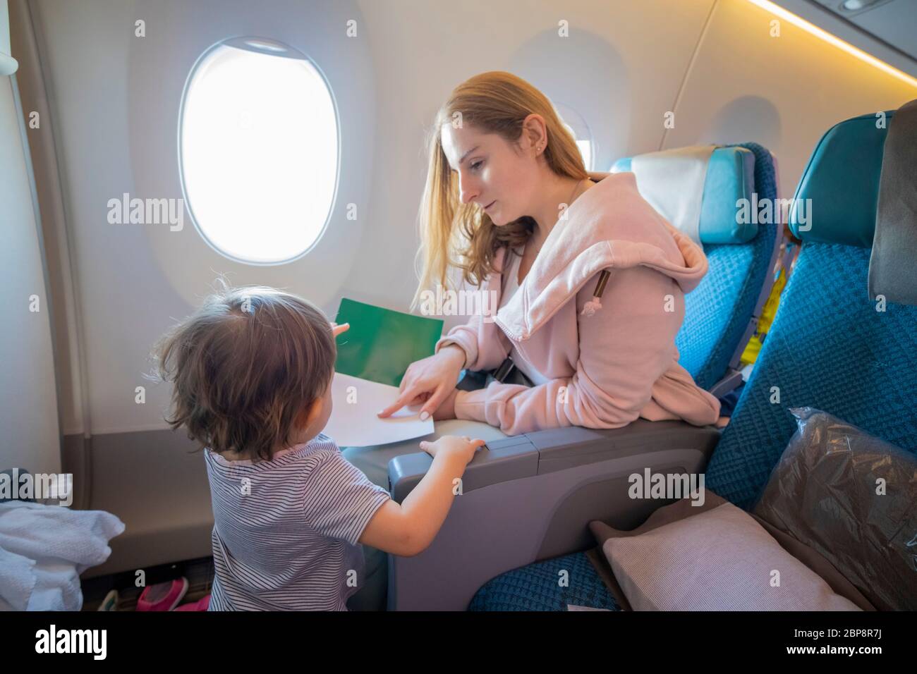 Eine junge schöne Mutter sitzt in einem Flugzeugsessel zeigt mit Finger eine Seite in einem Buch zu ihrem kleinen süßen Kleinkind, die vor ihr steht Stockfoto