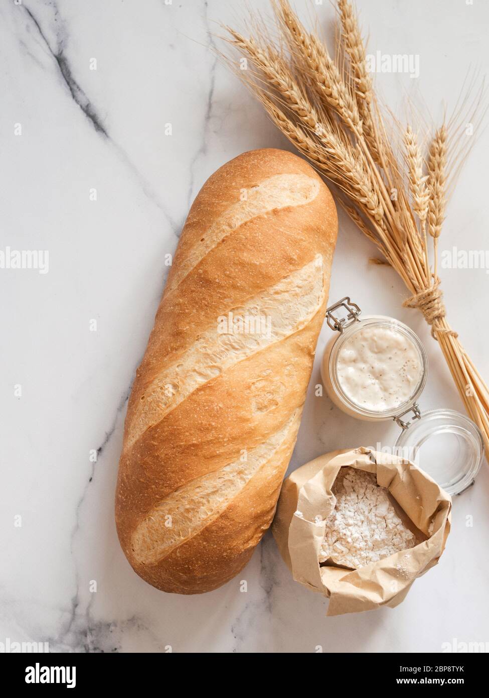 Britisches Weißblüher oder europäischer Sauerteig Baton-Brot auf weißem Marmorgrund. Frisches Brot und Glas mit Sauerteig Starter, Mehl in Papiertüte, Ohren. Draufsicht. Kopierbereich. Vertikal Stockfoto