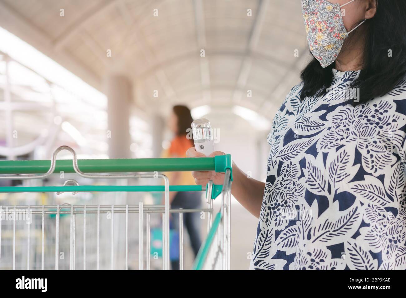 Frau tragen Maske mit Alkohol Nano-Sprühnebel antiseptische Reinigung auf Warenkorb Wagen Griff Schutz während Coronavirus Pandemie Covid-19. W Stockfoto