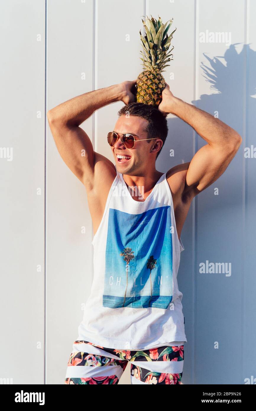 Gerne trendy Mann in Sonnenbrille hält eine Ananas auf dem Kopf, und das Lächeln, das Tragen von stilvollen Leibchen und Hosen. Im Freien. Stockfoto