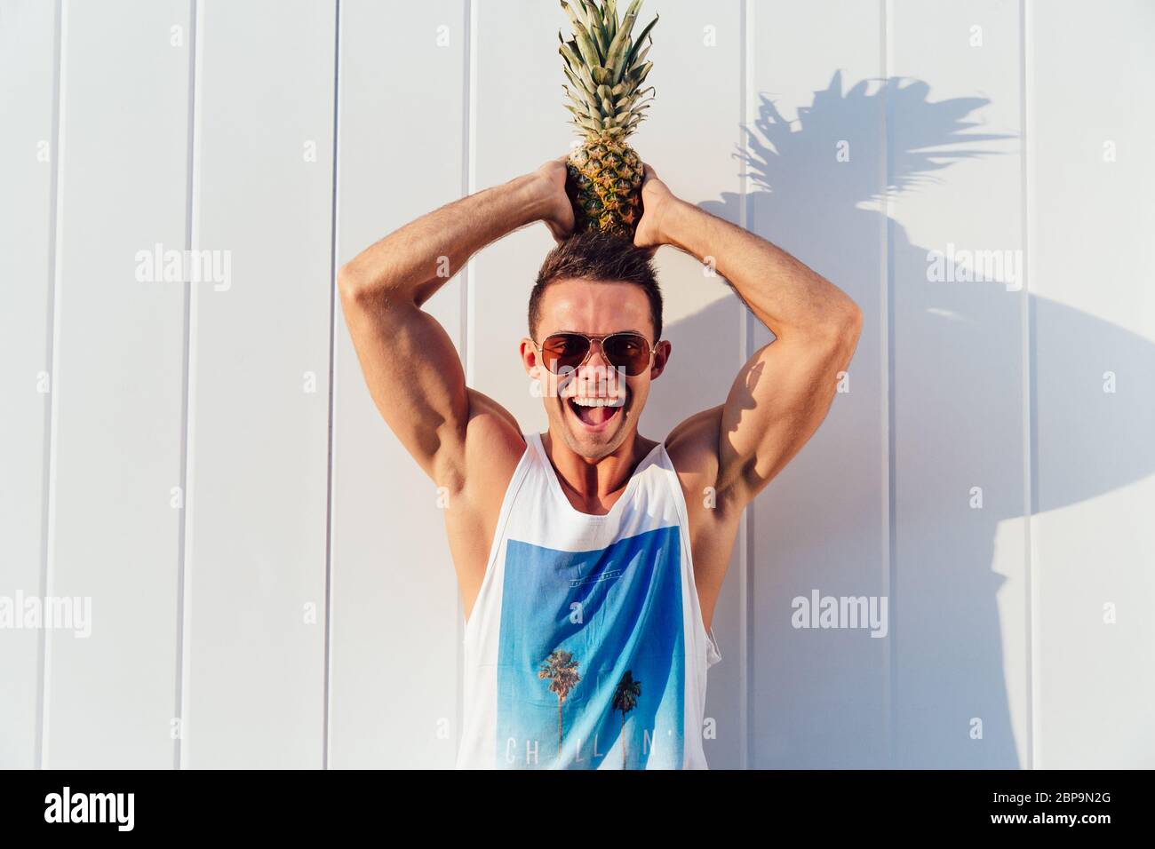 Freude schöner Mann in Sonnenbrille hält eine Ananas auf dem Kopf, in die Kamera schauen und lächeln, das Tragen von stilvollen singlet. Im Freien. Stockfoto