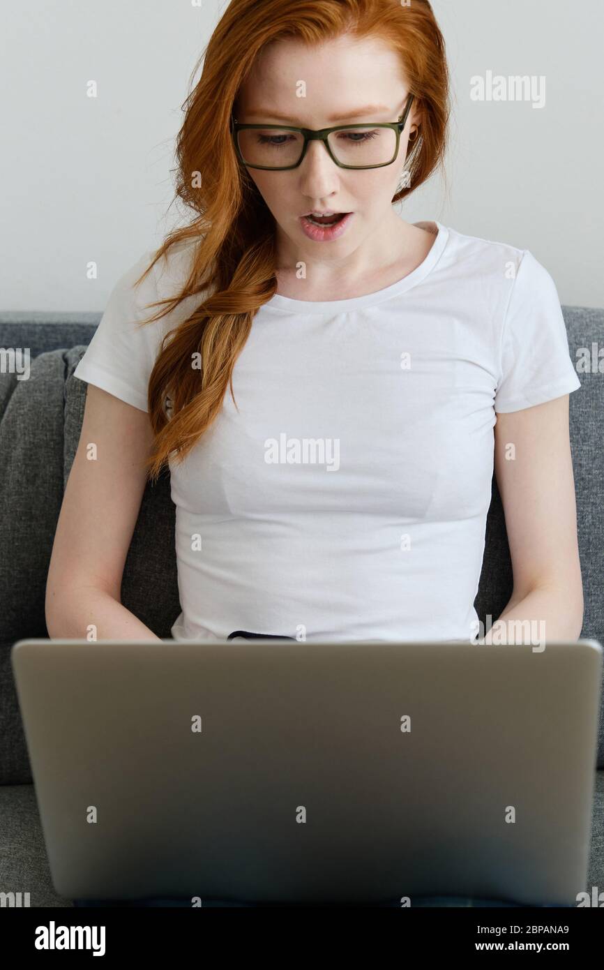 Ein rothaariges Mädchen sitzt auf einem Sofa in einem weißen T-Shirt und Brille, überrascht, öffnet ihren Mund, tippt auf einem Laptop Stockfoto