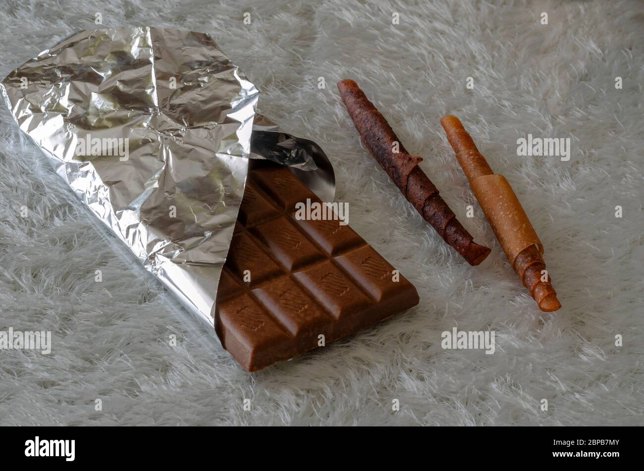 Eine Tafel mit offener Schokolade und zwei Stück Fruchtbonbons auf einem weißen, flauschigen Hintergrund. Schokolade in Folie und Süßigkeiten in einer Rolle verpackt. Natürlich gesund Stockfoto