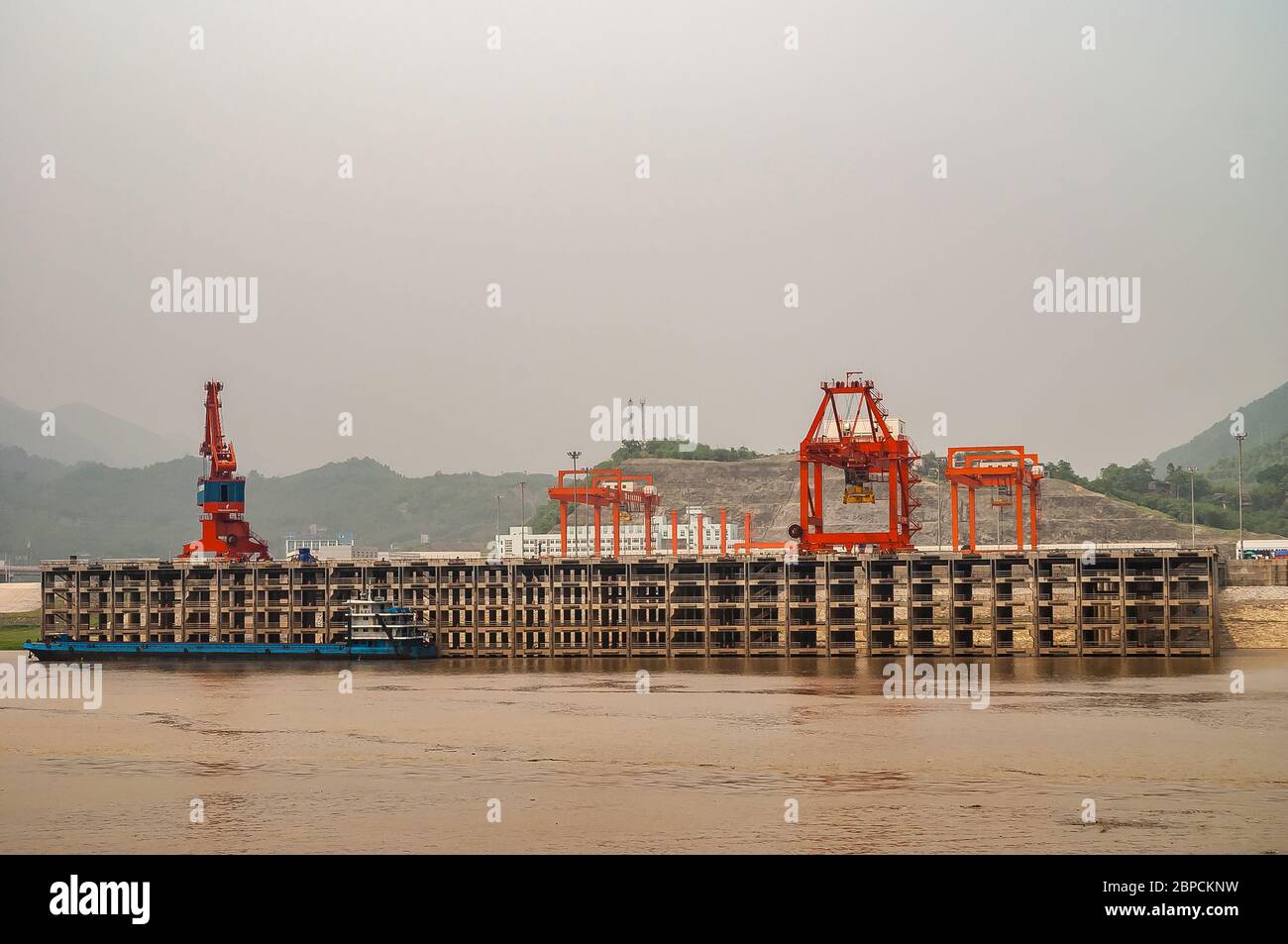 Huangqikou, Chongqing, China - 8. Mai 2010: Jangtse River. Landschaft von Rötelkräne auf mehrstufigen Hafenquai hinter braunem Wasser. Barge angedockt, Grün f Stockfoto