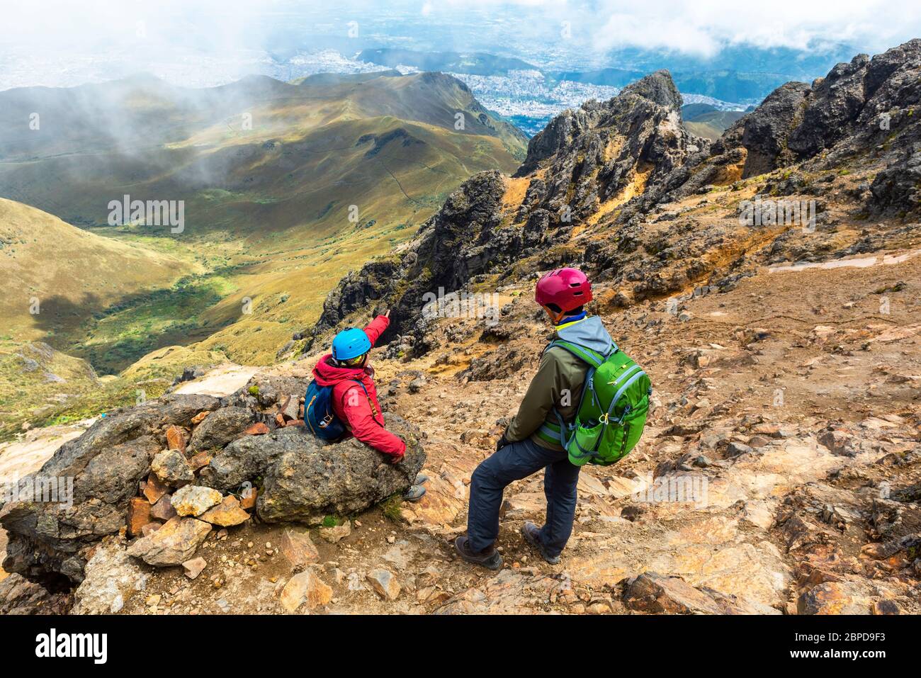 Zwei junge Backpacker mit Schutzhelm entlang der Rucu Pichincha Vulkanwanderung und mit dem Luftblick über Quito, Anden, Ecuador. Stockfoto